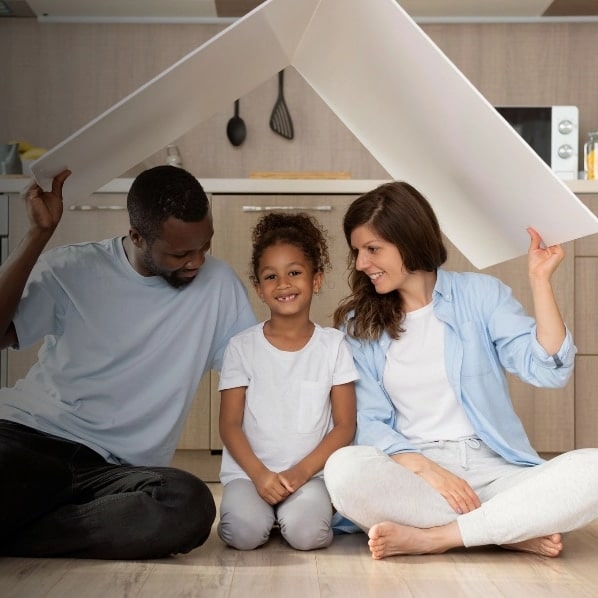 A man, a woman, and a young girl sit on a floor, smiling, holding a roof-shaped board above their heads, symbolizing a happy family at home.